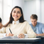 Modern Education Concept. Portrait of smiling female student sitting at desk in classroom at university, taking test or writing notes in her notebook, casual teen looking and posing at camera